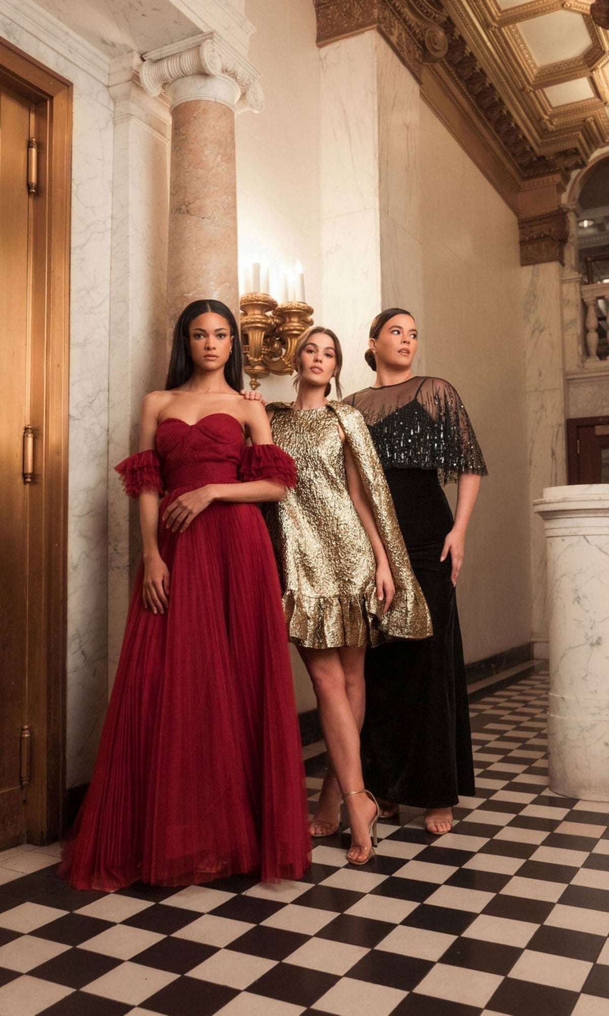 Three women stand in an elegant hallway with marble columns and a black and white checkered floor. One wears the Long Plus-Size Formal Dress: Mac Duggal 9228—a stunning black velvet gown with an embellished sheer cape.